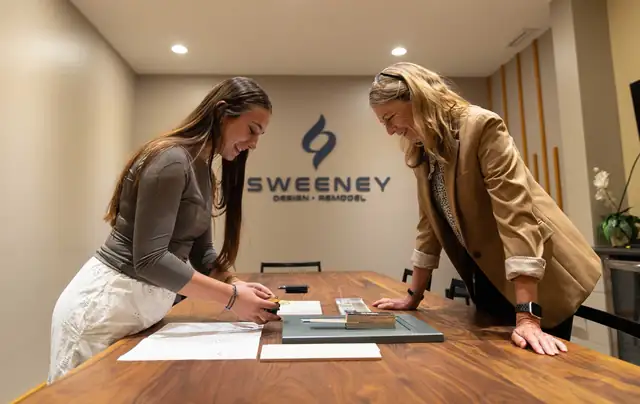 Two Sweeney designers laying out samples on a walnut table in front of the Sweeney Design Remodel logo wall